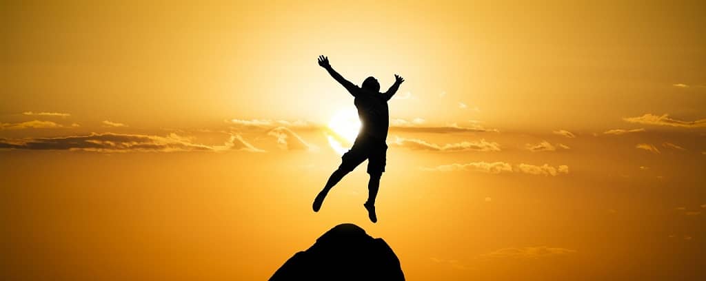 Person leaping into the air from a mountain peak, arms and legs outstretched while draped in a sunset.