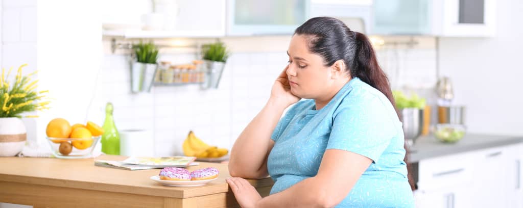 Photo of overweight person sitting at table looking sadly at plate of iced doughnuts
