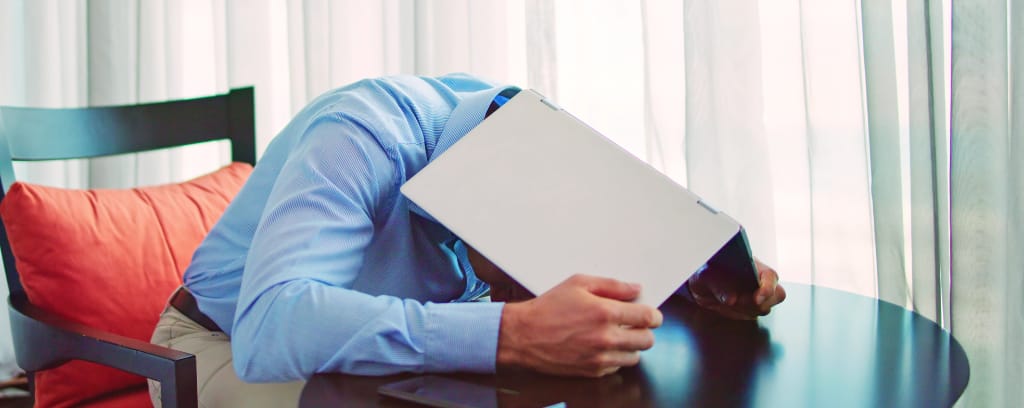 Photo of embarrassed worker hiding his head under an opened laptop turned upside down on a table.