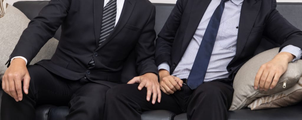 Two employees sit next to each other as one places a hand on the other's thigh